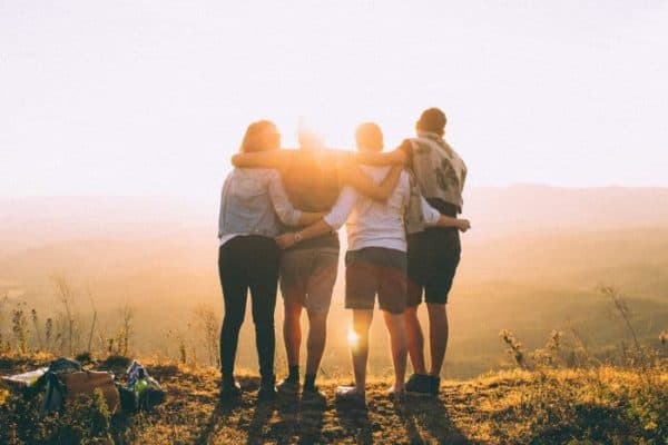 Rear view of four friends with their arms around each other facing the sunset - friends travel lifelong learning