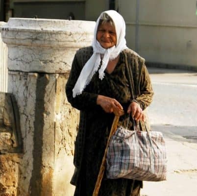 Elderly Roma Woman with weathered skin. She wears a white headscarf and long brown dress. She's carrying a wooden cane and a plaid tote bag in Sarajevo, Bosnia