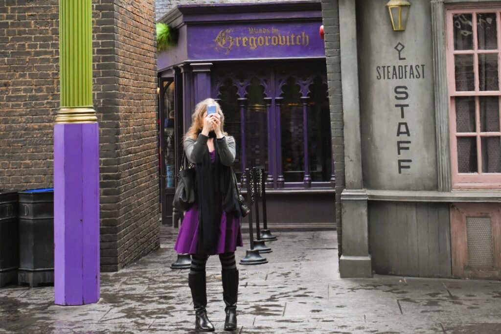 A young woman in a purple dress, striped tights and black boots stands taking a picture with a cell phone hiding her face. She is standing next to a purple and bright green lamp post in Diagon Alley at Universal Orlando Harry Potter World