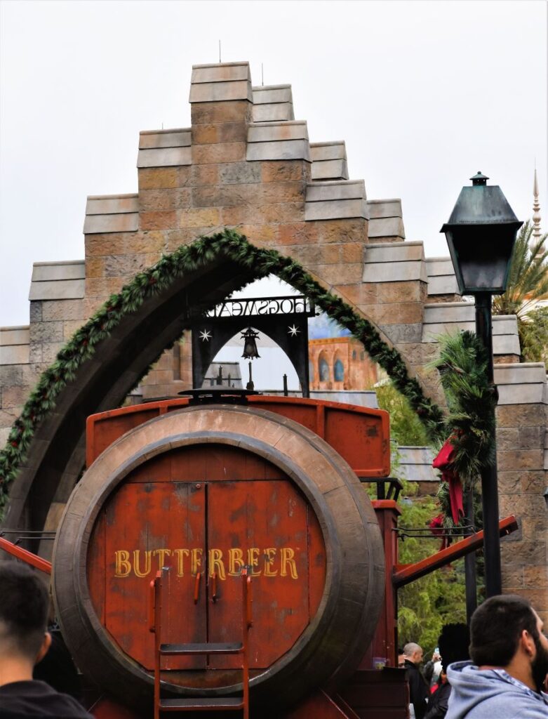 A large wooden barrel with the words "Butter Beer" sits on top of a rustic cart. The cart is passing under a brick arch with the words "Hogsmeade".