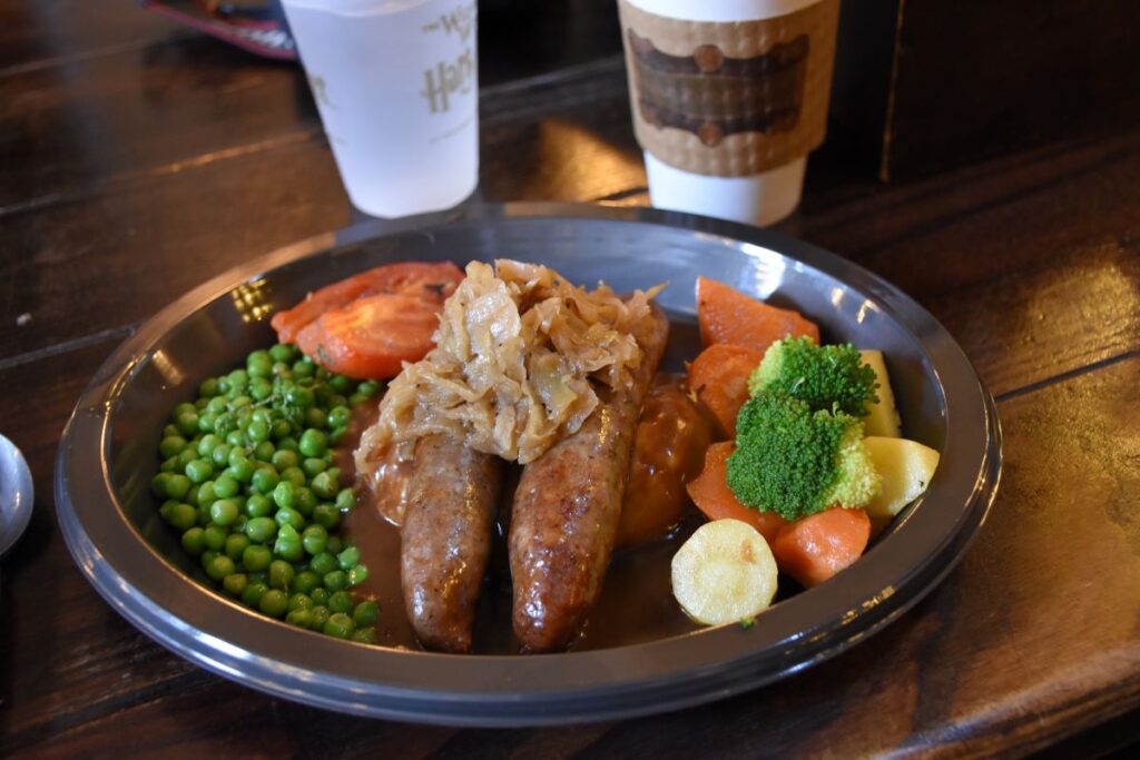 A plate of Harry Potter food at Universal Orlando - bangers and mash with green peas, carrots, broccoli and a stewed tomoto on a metal plate sitting on a wood table