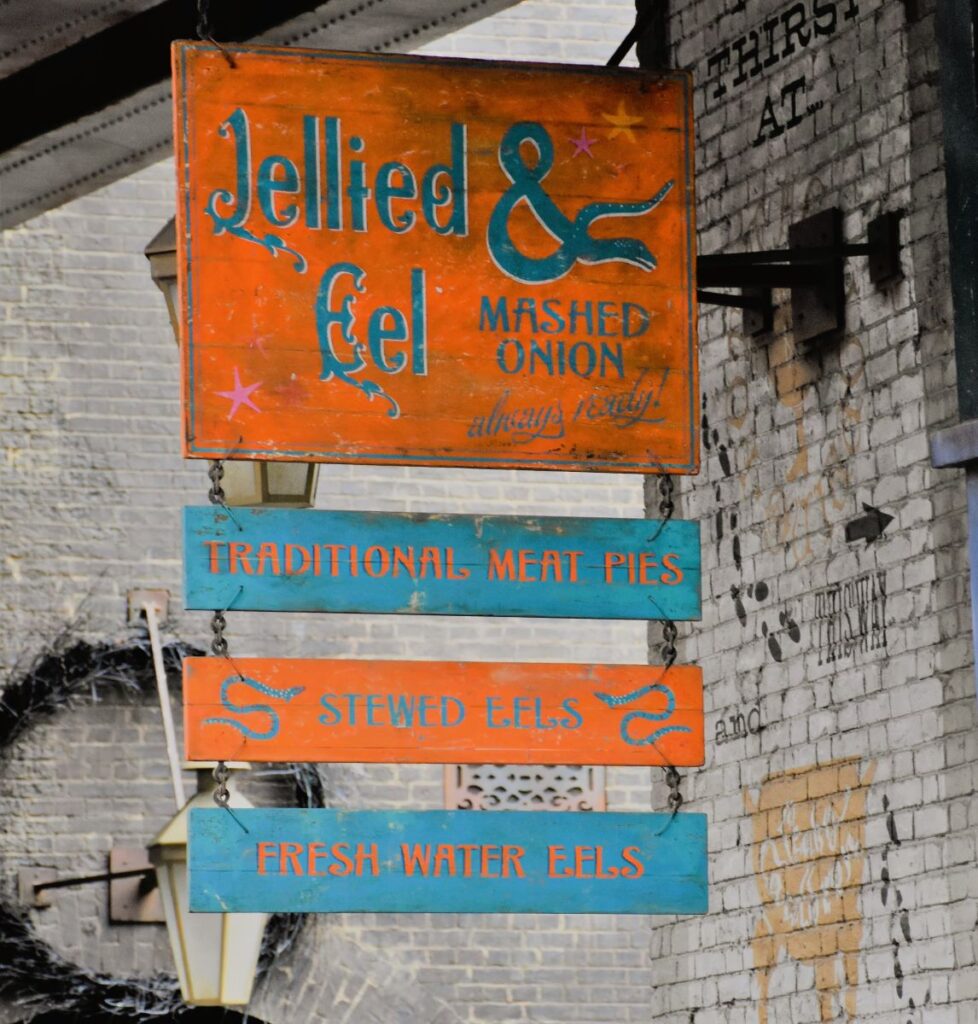 An orange and blue wooden sign with 4 boards hanging in succession says "Jellied Eel and Mashed Onion - Always ready", "Traditional Meat Pies", "Stewed Eels" and "Fresh Water Eels" Harry Potter foods