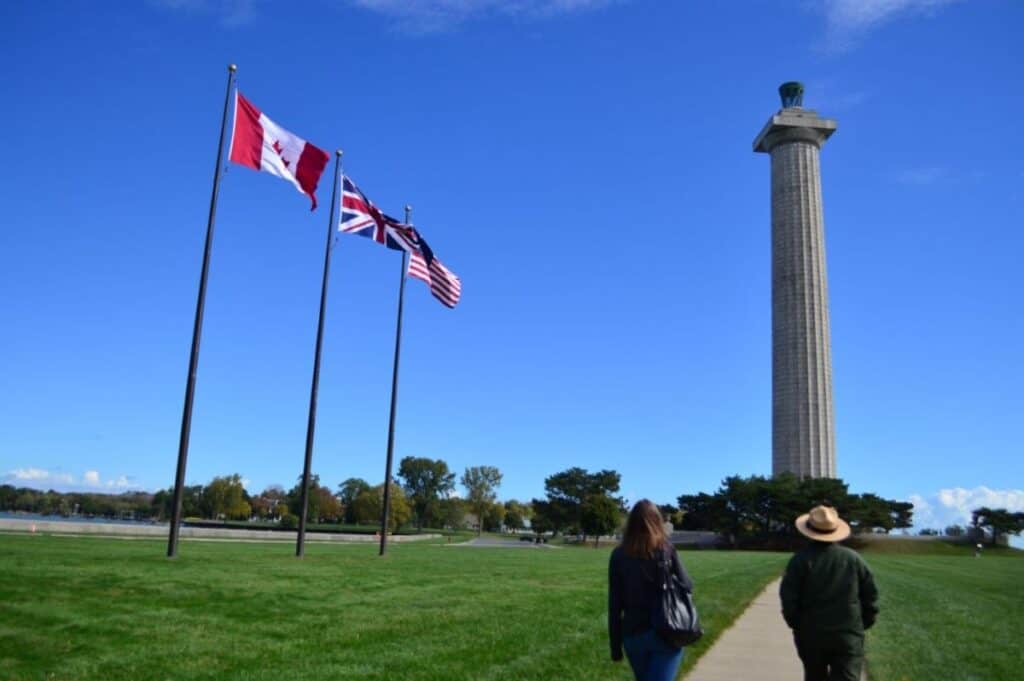 Park ranger and woman seen from the back walking toward Perry's Monument in Ohio. It's a 352 foot tall gray round column rising in the air. The Canada, Great Britain and U.S. flags fly nearby.
