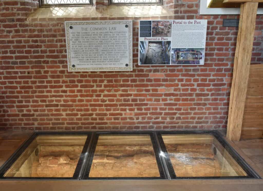Glass floor covers archeological view of church building foundation from the 1600s in Jamestowne Virginia. A plaque above explains more details. Historical travel in the U.S.