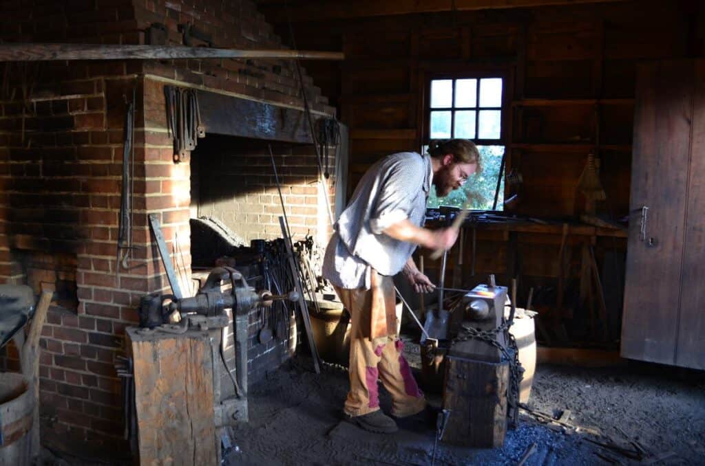 Blacksmith striking hot metal rod with a metal hammer. He's standing in front of a huge fireplace with blacksmithing tools around him at George Washington's Mount Vernon. Historical travel in the U.S. 