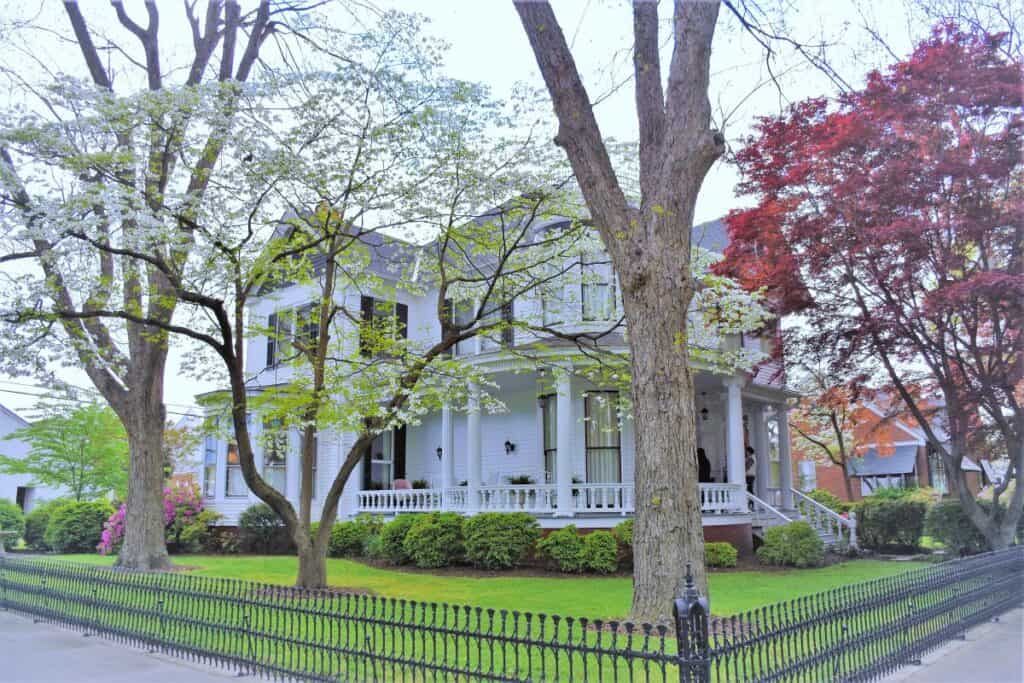 A stately white historic home with black shutters and a wrap around porch in West Point Virginia. Several trees and a decorative wrought iron fence surround the house.