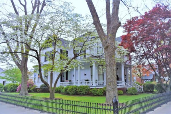 A stately white historic home with black shutters and a wrap around porch in West Point Virginia. Several trees and a decorative wrought iron fence surround the house.