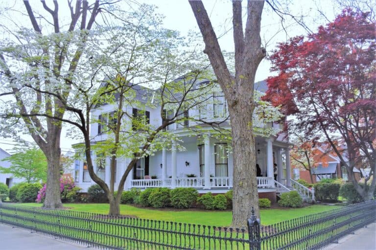 A stately white historic home with black shutters and a wrap around porch in West Point Virginia. Several trees and a decorative wrought iron fence surround the house.