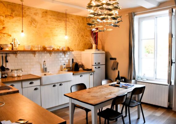 A beautiful kitchen with a rustic stone wall, white beachy cabinets and a wooden table for 4 in the center. A beautiful light fixture with silver fish cutouts hangs above the table and a window on the far walls allows light to stream in. Port-en-Bessin - lodging near Normandy Beach, France