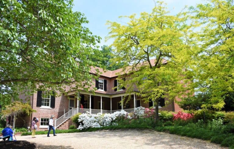 Historic brick two-story home with black shutters and a large front porch. Green trees and white flowers surround the home and several people are seen walking around and sitting near the house near Fredericksburg Virginia