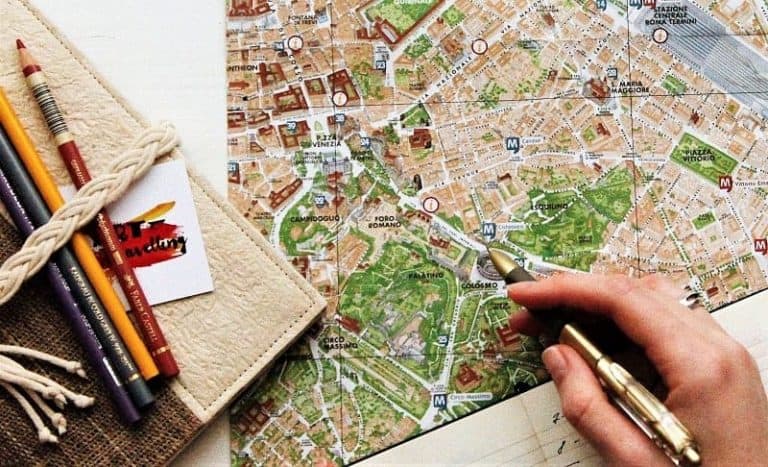 Hand holding a pen over a map making travel plans. Additional colored pencils sit off to the side.