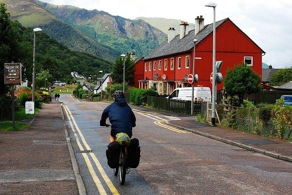 Person riding a bike down a highway through a small village in Glencoe, Scotland