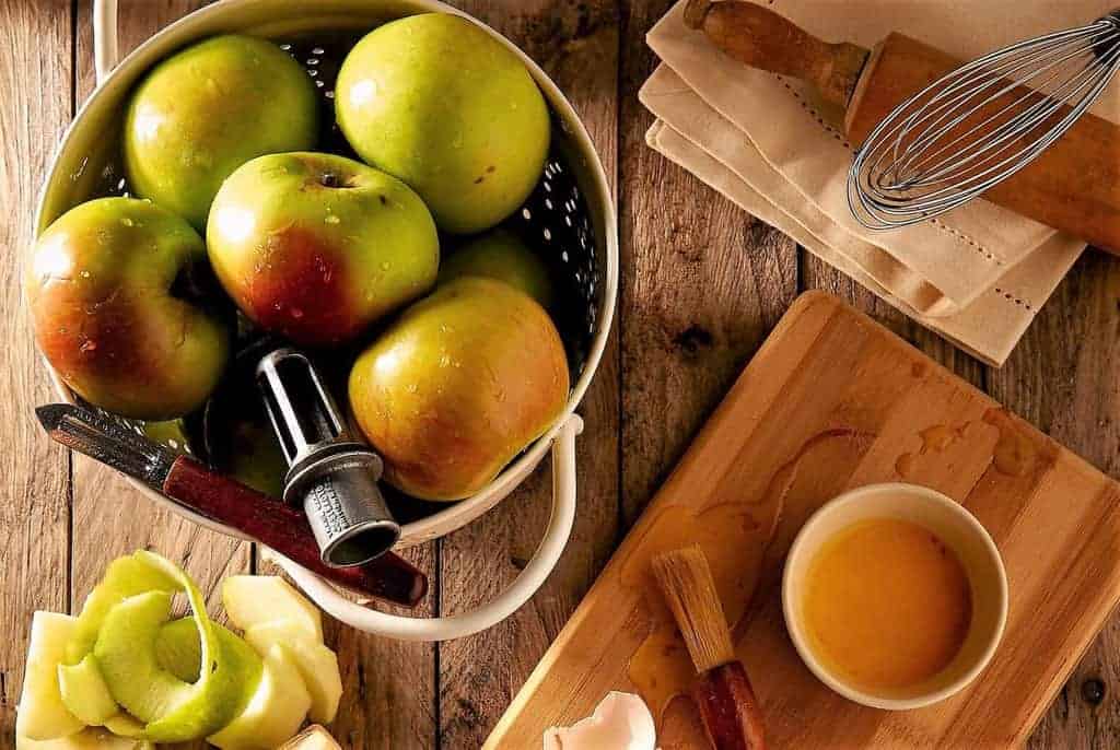 A colander with green and red apples, an apple peeler with several apple skins next to a wooden cutting board. rolling pin and whisk. Normandy France is known for apple cider, Calvados, and apple brandy