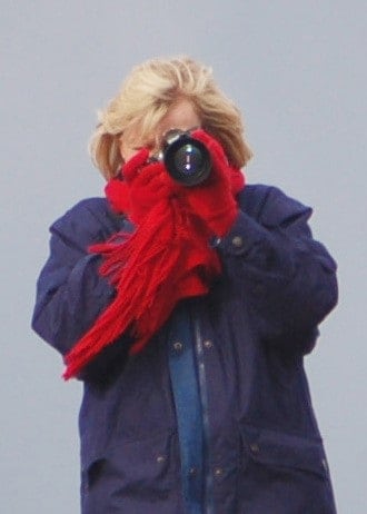 Woman with blonde hair holding a DSLR camera up to her face to take a picture. She has on red gloves and a red scarf. Nancy Hann Lifelong Learner