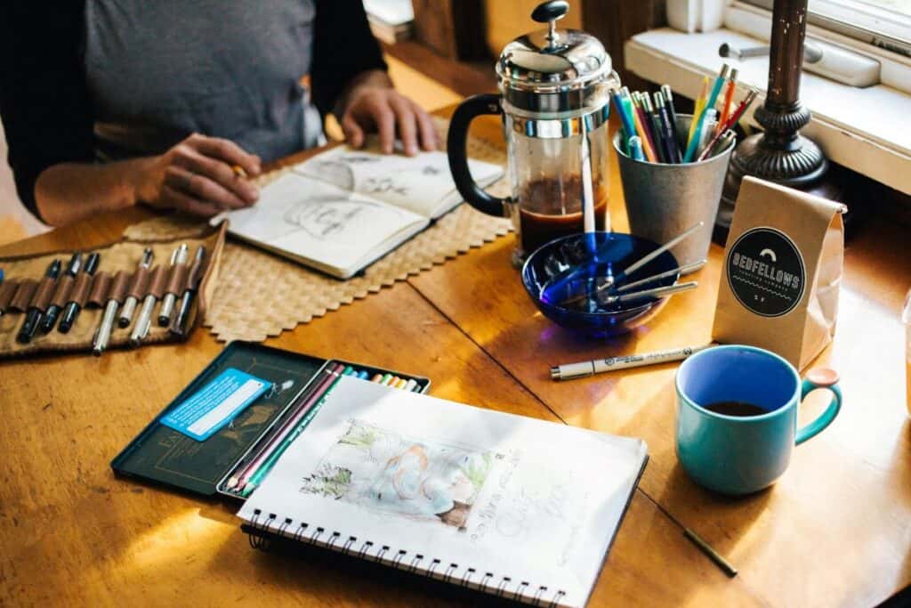 A person sits at a wooden table next to a window, working on a sketch in a notebook. On the table, there is another open notebook with a watercolor painting of flowers, a blue mug, and an assortment of drawing tools including pencils, brushes, and markers. A French press coffee maker and a bag labeled “COFFEE” are also present at the artistic workspace setup.