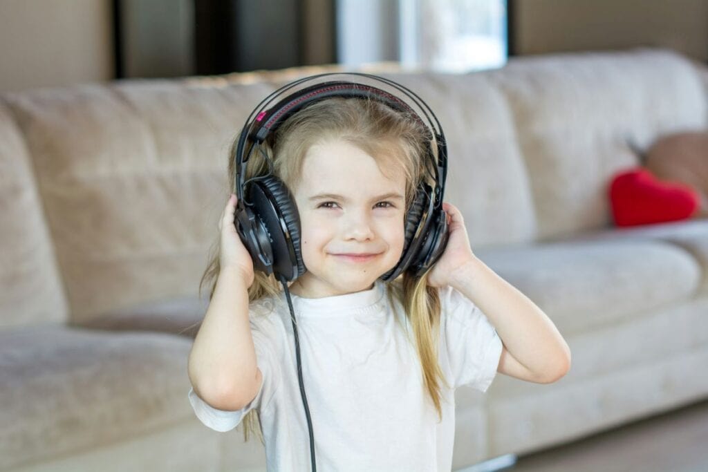 A smiling child in a bright room stands in front of a beige couch, holding large black headphones over their ears. The girl is listening to music, which is one of the fun things to do when you’re bored.