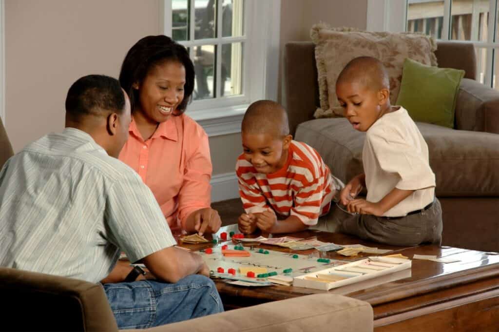 A family of two adults and two boys is seated around a coffee table engaged in playing a board game in a cozy living room. This activity is one of many fun things to do when you’re bored.