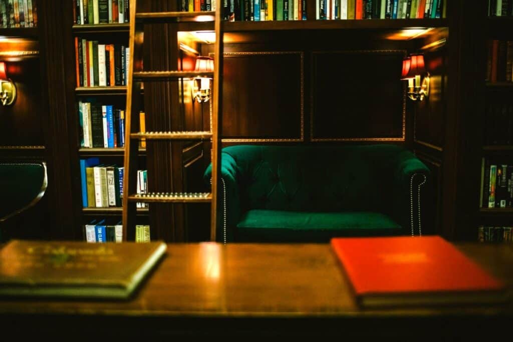 A cozy library setting with a dark wood bookshelf filled with various books. In the center, a plush green sofa invites reading and relaxation. The foreground shows a table with a focused view of an orange book, suggesting a quiet and studious atmosphere in the library. A wooden ladder leans against the bookshelf, adding a classic touch and providing access to the higher shelves.