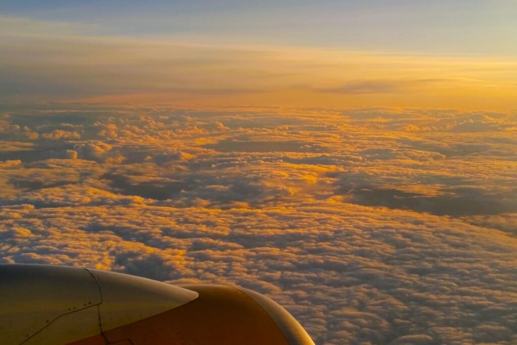 The view from an airplane window showing the edge of the engine and a cloud-covered sky from above the clouds with the yellow and orange of the sunset shining on the top of the clouds. 