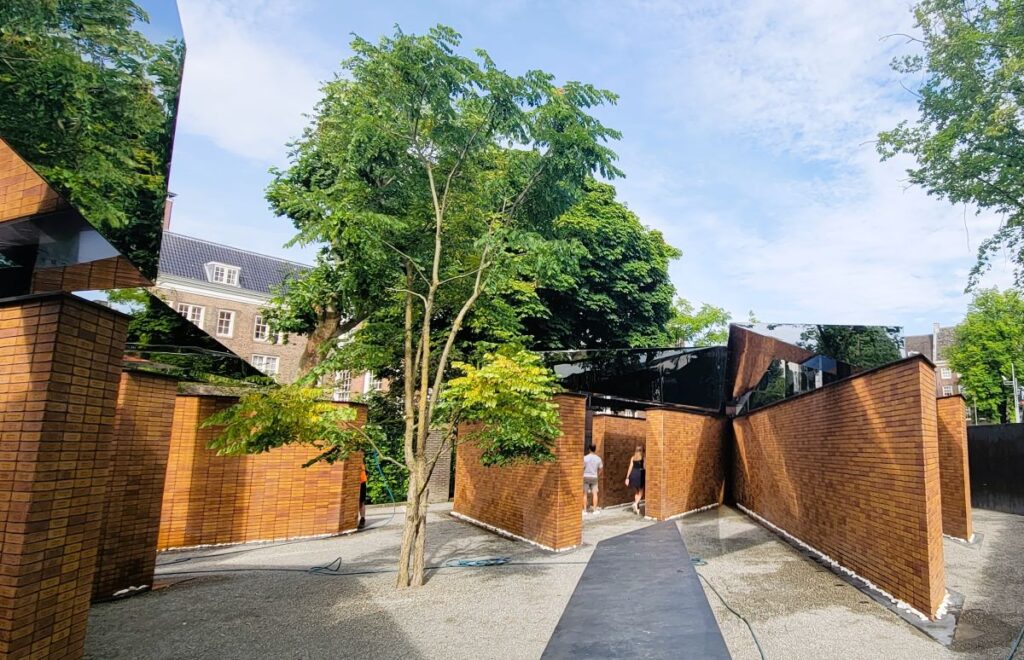 Walls of orange brick with a name inscribed on each one and walls of mirrors above the brick. The names represents 102,000 Jewish victims and 220 Sinti and Roma victims who lost their lives in the Netherlands during the Holocaust of WWII. The memorial is designed to commemorate the victims individually and collectively.