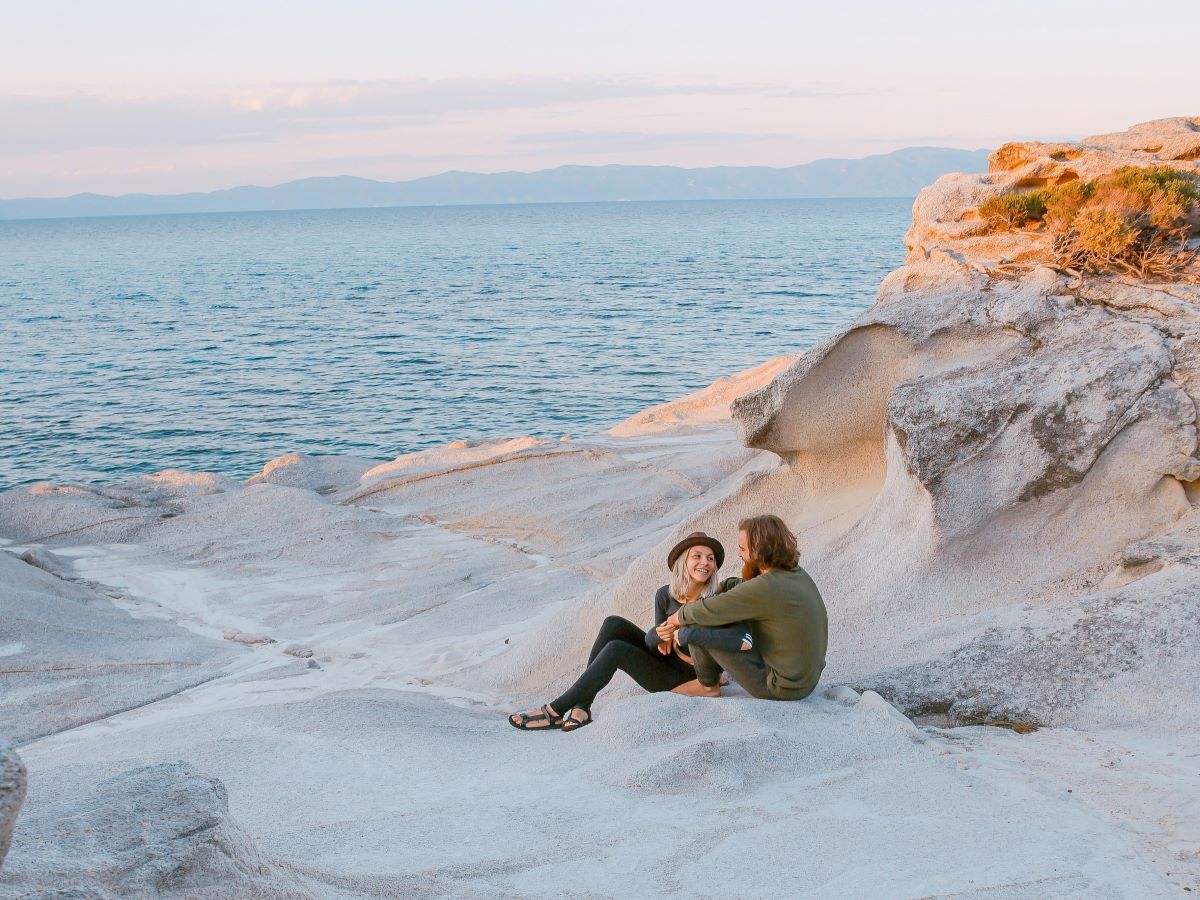 A young happy couple looks at each other while sitting on a white sandy and rocky beach next to calm blue water with hills in the distance.