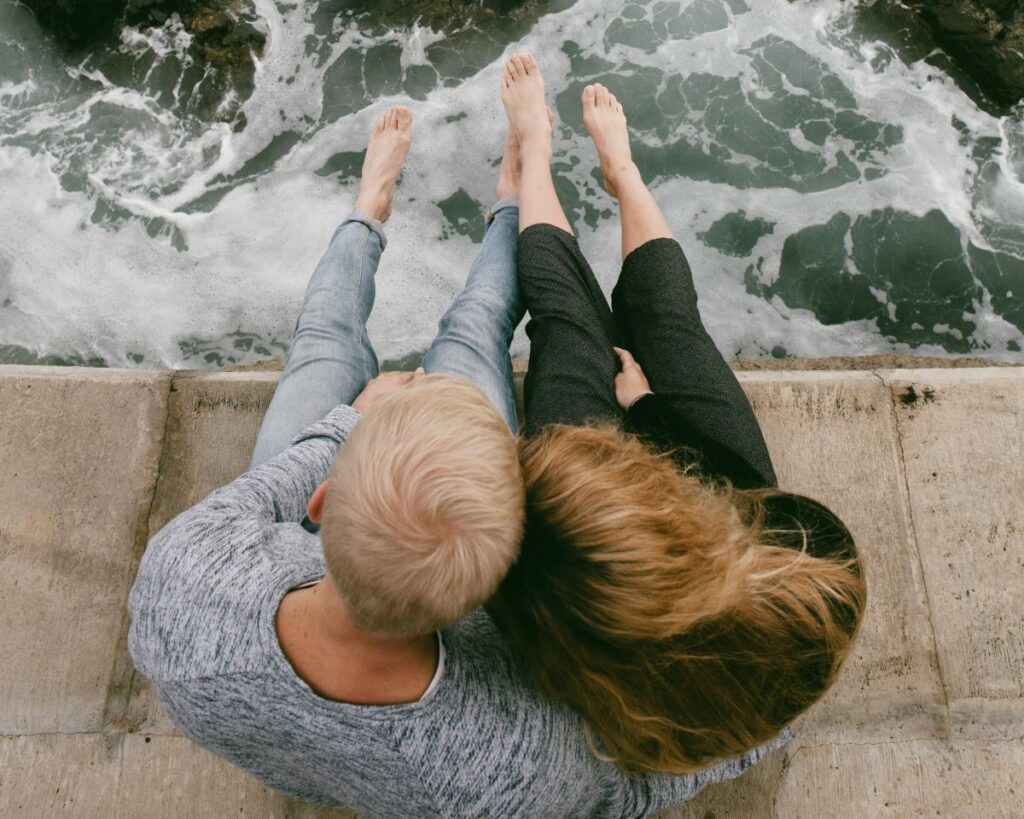 An overhead view of a romantic couple snuggling close together on a concrete pier with their bare feet dangling above the ocean waves below.