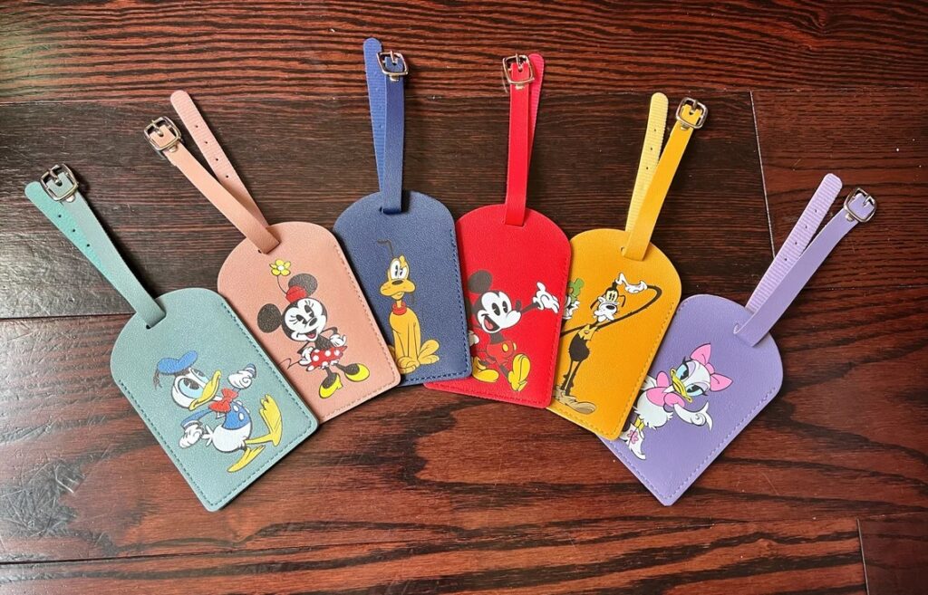 Six different leather luggage tags, each a different color with a different Disney character on the front. They each have a small strip of leather with a buckle to attach to the bag. 