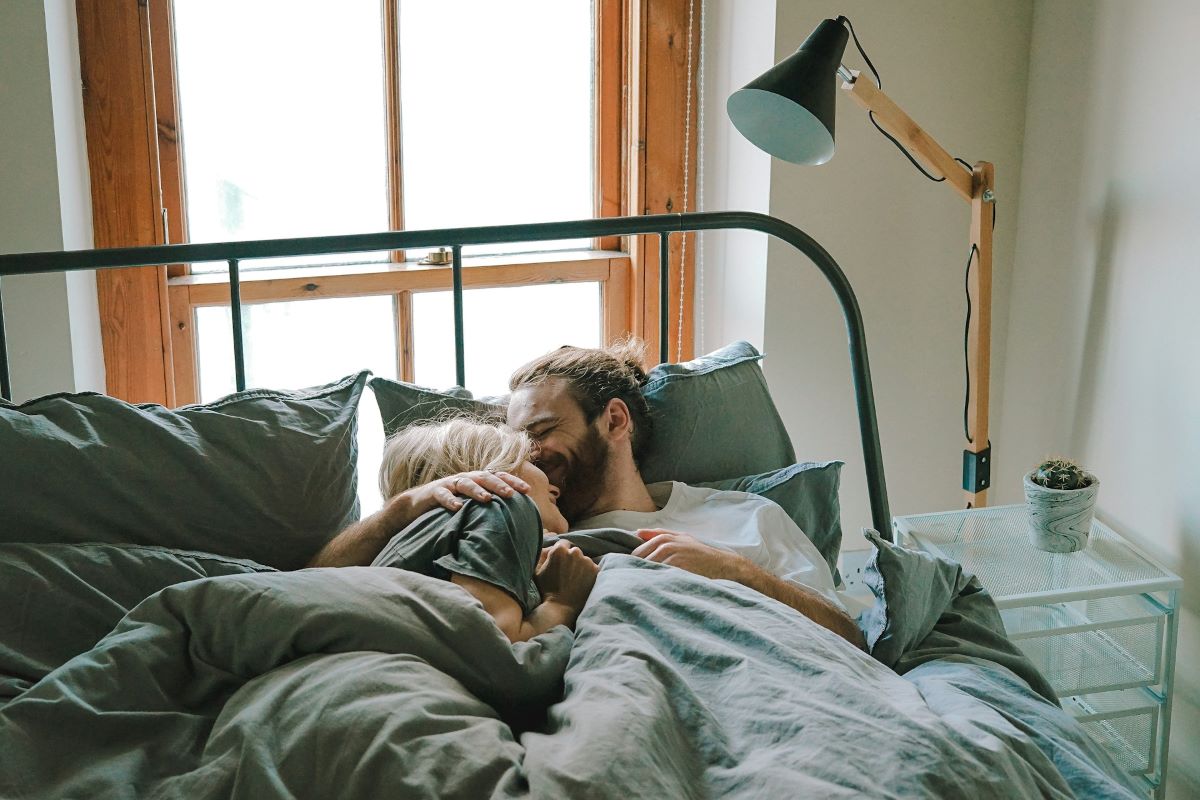 A young hopeless romantic couple snuggled in bed under sage green sheets, sharing a quiet, intimate moment by a large window with soft natural light streaming in.