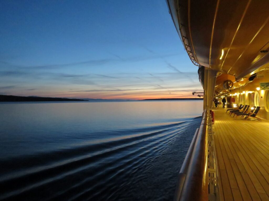 The deck of a cruise ship with deck chairs facing toward the water. The water is calm and the sunset can be seen in the distance.