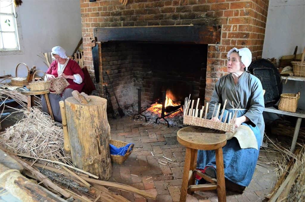 Two women dressed in Colonial clothes weaving baskets in front of a large fireplace