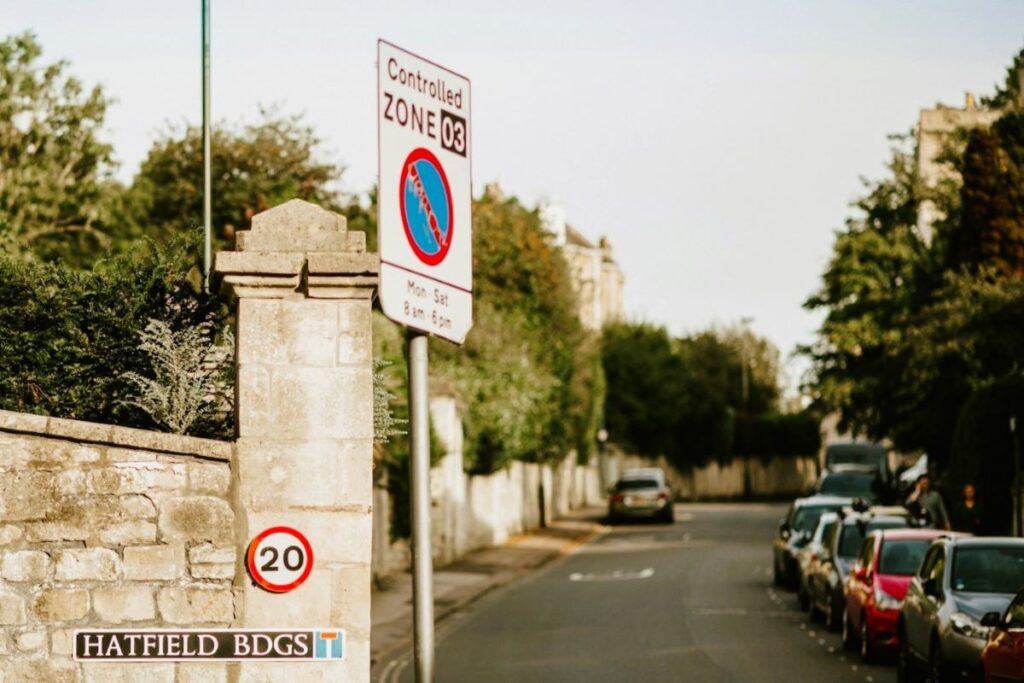 Residential street in Bath, England with cars parked along one side and a sign says "Controlled Zone 03", with a 20 KMH speed limit sign posted on a stone wall. Parking is one reason to consider  travelling to Bath England without a car.