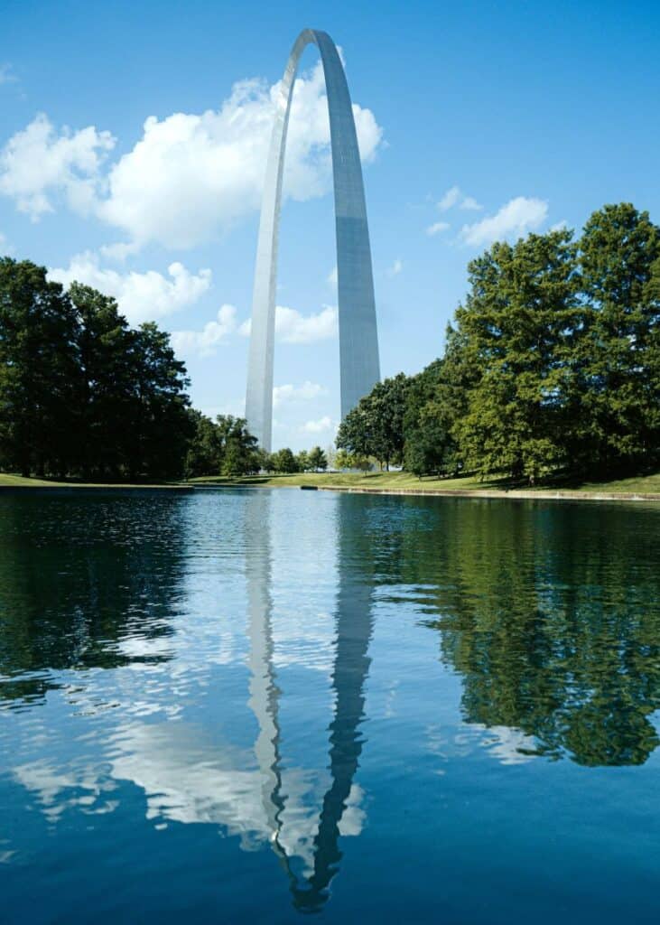 Giant metal arch in Saint Louis, Missouri Gateway Arch with trees on both sides and water in the foreground with reflection of the arch