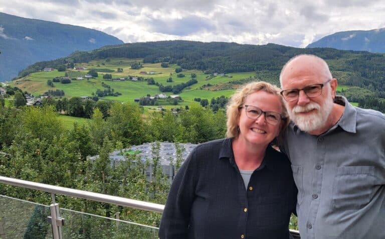 Man with glasses and white beard and woman with glasses and short blonde hair in Ulvik, Norway. Green hillsides speckled with small homes in the background.