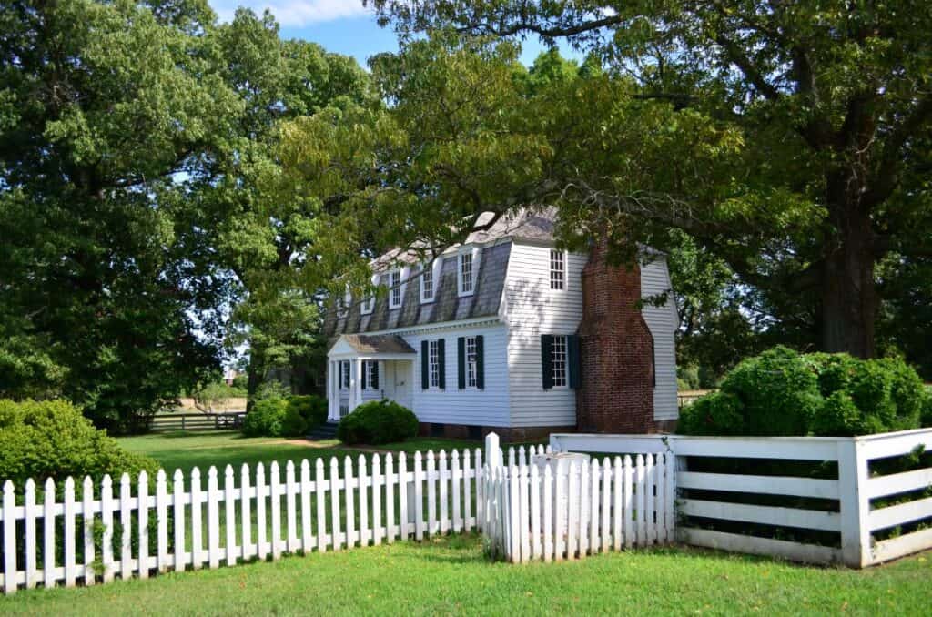 The Moore House where the negotiations of surrender for the Revolutionary War took place in Yorktown, VA 1781. A white colonial house with 9 windows on the front and a red brick chimney on the right side. A white picket fence surrounds the house. Green grass and shady trees are all around the house. 