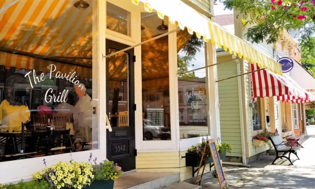 Historic storefronts on quaint main street in Vermilion Ohio. The Pavilion Grill has colorful striped awnings and large windows. Flower boxes filled with fresh yellow flowers sit in front of each store.