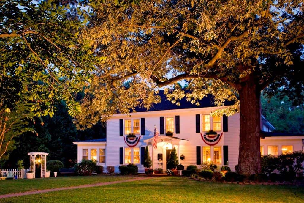A white 2-story historic home with black shutters on the windows lit up at night. Large trees overlook the home.  Williamsburg, VA