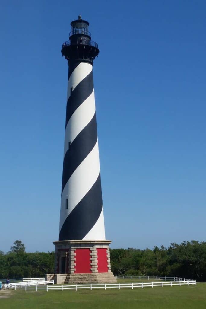 Black and white spiral painted lighthouse with red and brick base surrounded by a white rail fence. Cape Hatteras North Carolina