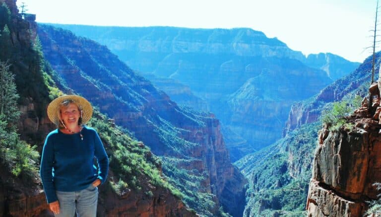 Woman with blonde hair, wide-brimmed straw hat and teal long sleeve shirt smiling at the camera. The Grand Canyon cavern is in the background in shades of blue and green. Nancy Hann traveling with purpose.