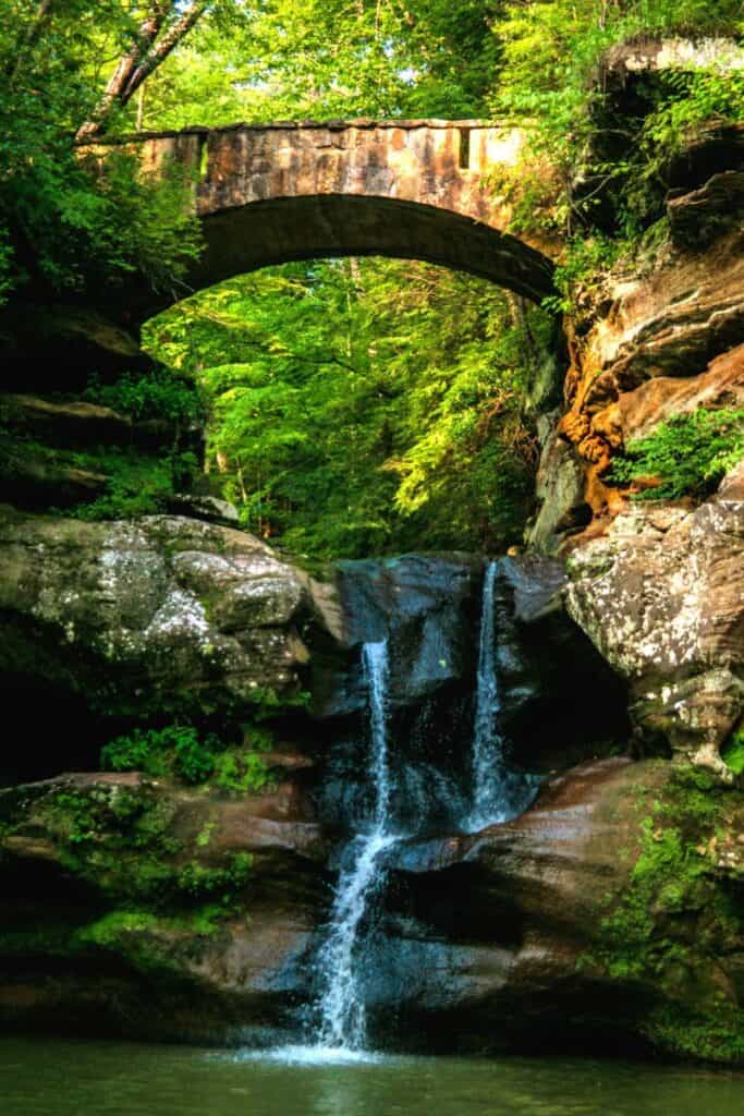 Lush green forest with arched bridge above and trickling waterfall lined with boulders below. Hocking Hills Ohio