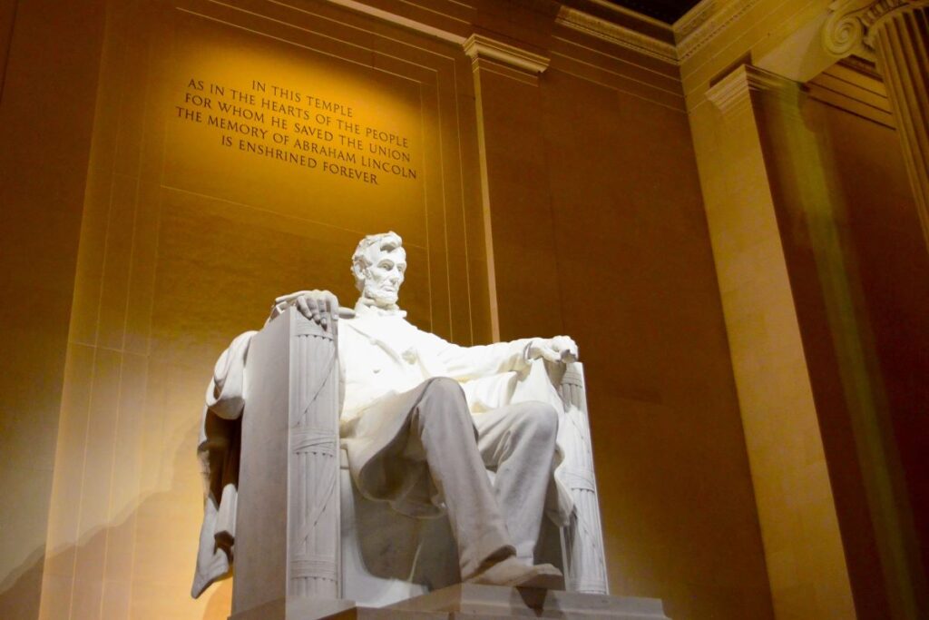 Abraham Lincoln marble statue inside the Lincoln Memorial building in Washington DC - A thin man with a short beard sits in a suit in a large chair with his arms resting on the arms of the chair. Engraved on the wall above his head are the words, "In this temple as in the hearts of the people for whom he saved the Union, the memory of Abraham Lincoln is enshrined forever."