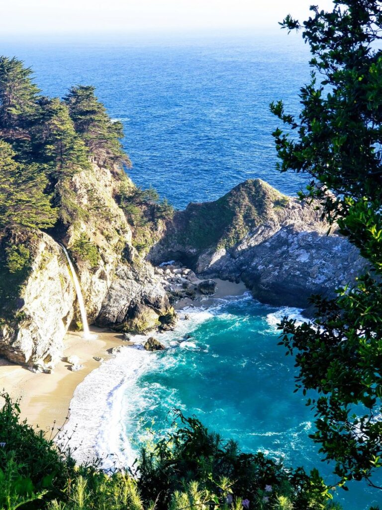 Julia Pfeiffer Burns State Park waterfall coming out of the coastal rocks and falling down to the beach on the California rocky coastline. Turquoise and blue ocean water can be seen washing up on the beach.