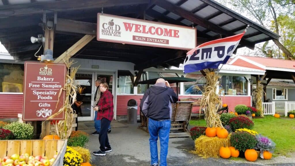 A man walks into a red building with fall plants in front of it. Bright orange pumpkins are stacked on top of straw bales and fall colored mums sit next to them. The sign says Cold Hollow Cider Mill - Welcome