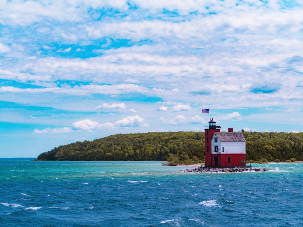 Round Island Lighthouse - A red and white building with lighthouse and American flag flying above. It sits on a small island of rocks in the turquoise blue waters of Lake Michigan near Mackinac Island. A forest of green trees is in the background with partly cloudy blue skies above.