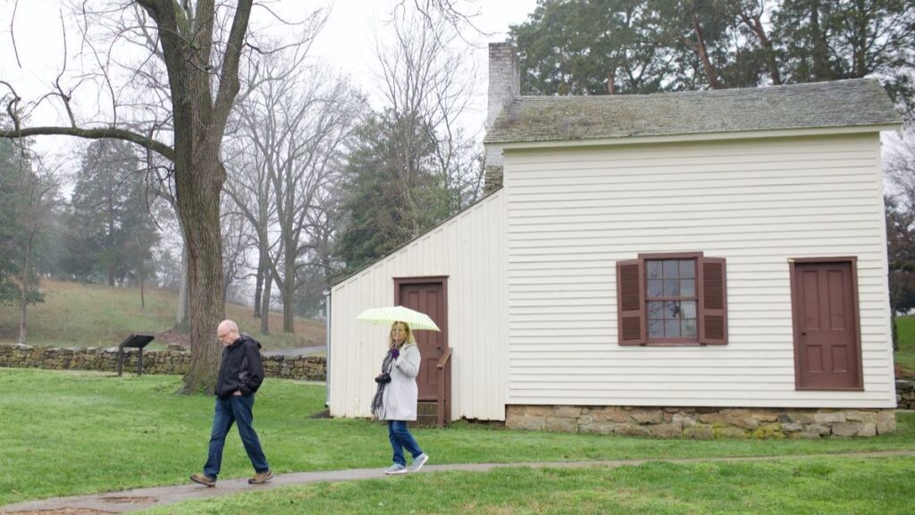 A man and woman walk in the rain. The woman is carrying an umbrella. There is a white historic wood home in the background. Fredericksburg, VA