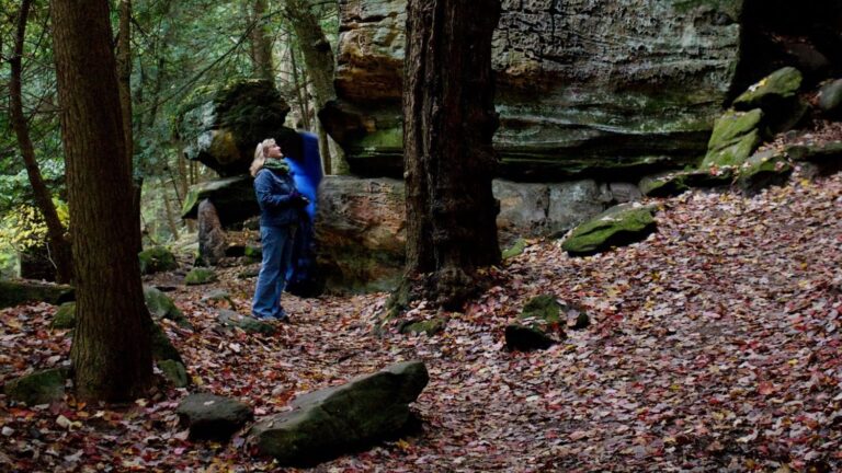 Woman with blonde hair and a denim jacket stands in a forest looking up at a tall moss-covered rock formation. There are brown fall leaves covering the ground. Cuyahoga Valley National Park, Ohio - Traveling with Purpose