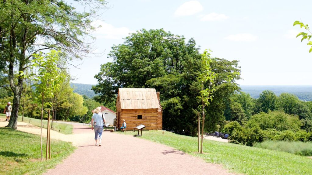 Woman with blonde hair carrying a camera walks away from the viewer, down a wide dirt path toward a small log cabin that depicts what enslaved quarters looked like at Thomas Jefferson's Monticello plantation in Virginia. There are trees behind the building and in the distance.