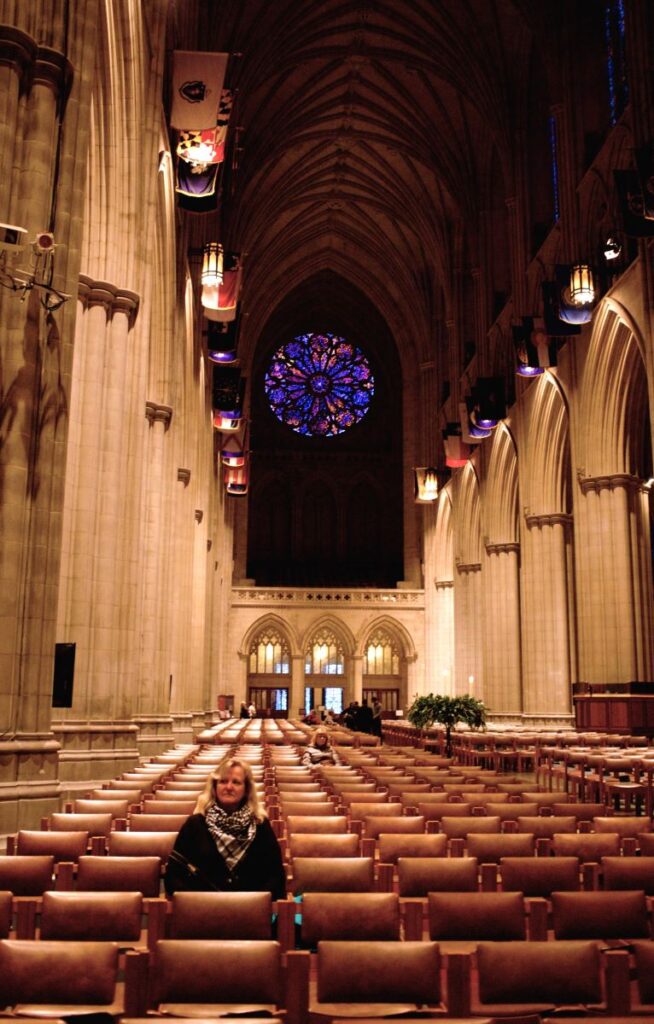 Blonde woman with black coat and black and white scarf sits in a row of chairs in a massive cathedral with soaring arched ceilings. A large round intricate stained glass window with shades of blue and red sits high on the wall behind her. National Cathedral in Washington DC