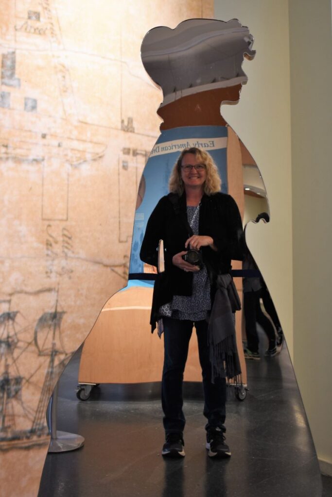 A woman stands smiling at her reflection and pointing her camera in front of a mirror that is shaped like a cutout silhouette of a woman in a long dress. Traveling with purpose - learning about history in Philadelphia PA.