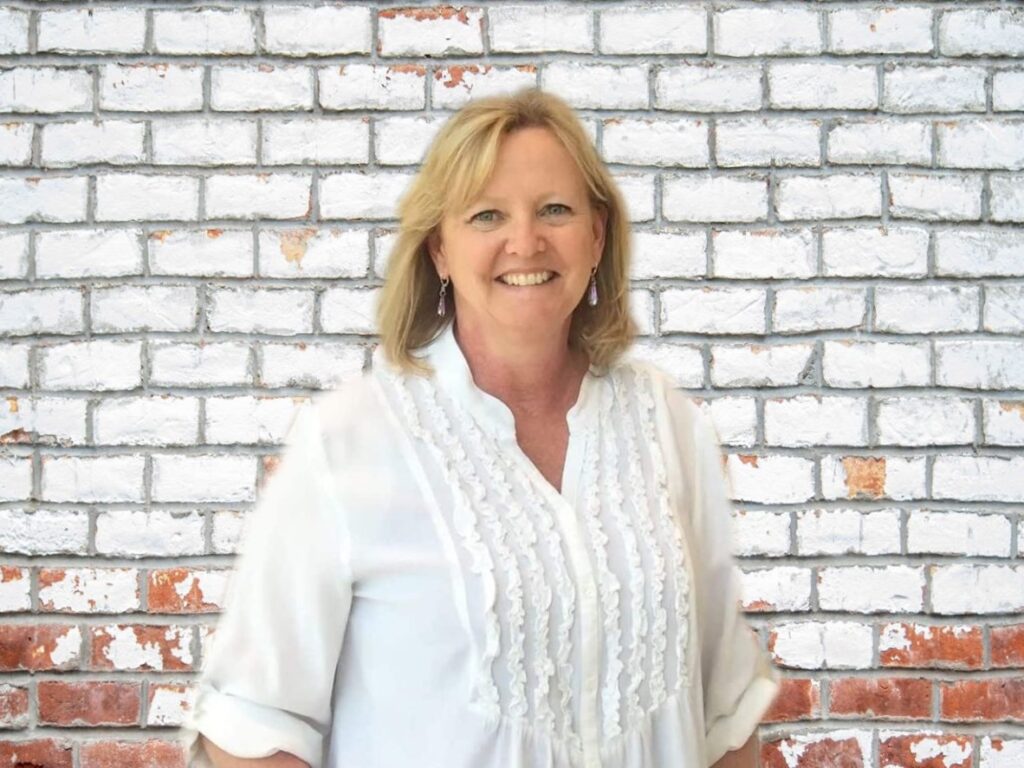 Woman with blonde hair smiling at the camera. She is standing in front of a rustic brick wall painted white. Nancy Hann - travel blogger