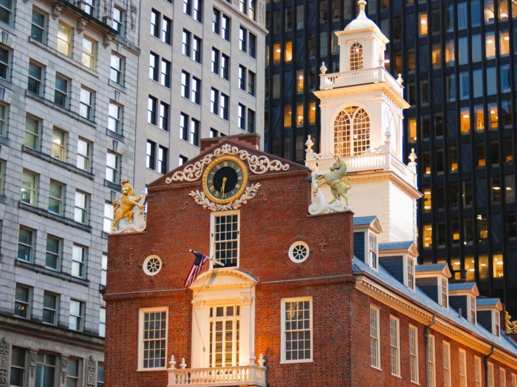 The Old State House in Boston, Massachusetts at dusk - A historic brick building surrounded by high rise buildings. There is a white door with a balcony on the front of the 2nd floor. A large clock sits at the peak of the building. A lion and unicorn statue stand on either side of the clock. A beautiful white tower rises above the main structure. This is the oldest public building in Boston.